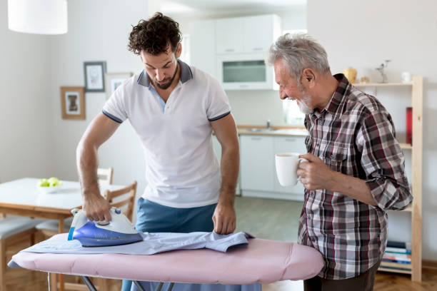 auxiliaire-de-vie-repassage Un auxiliaire de vie homme faisant le repassage avec un sénior à côté tous les deux discutants en souriant